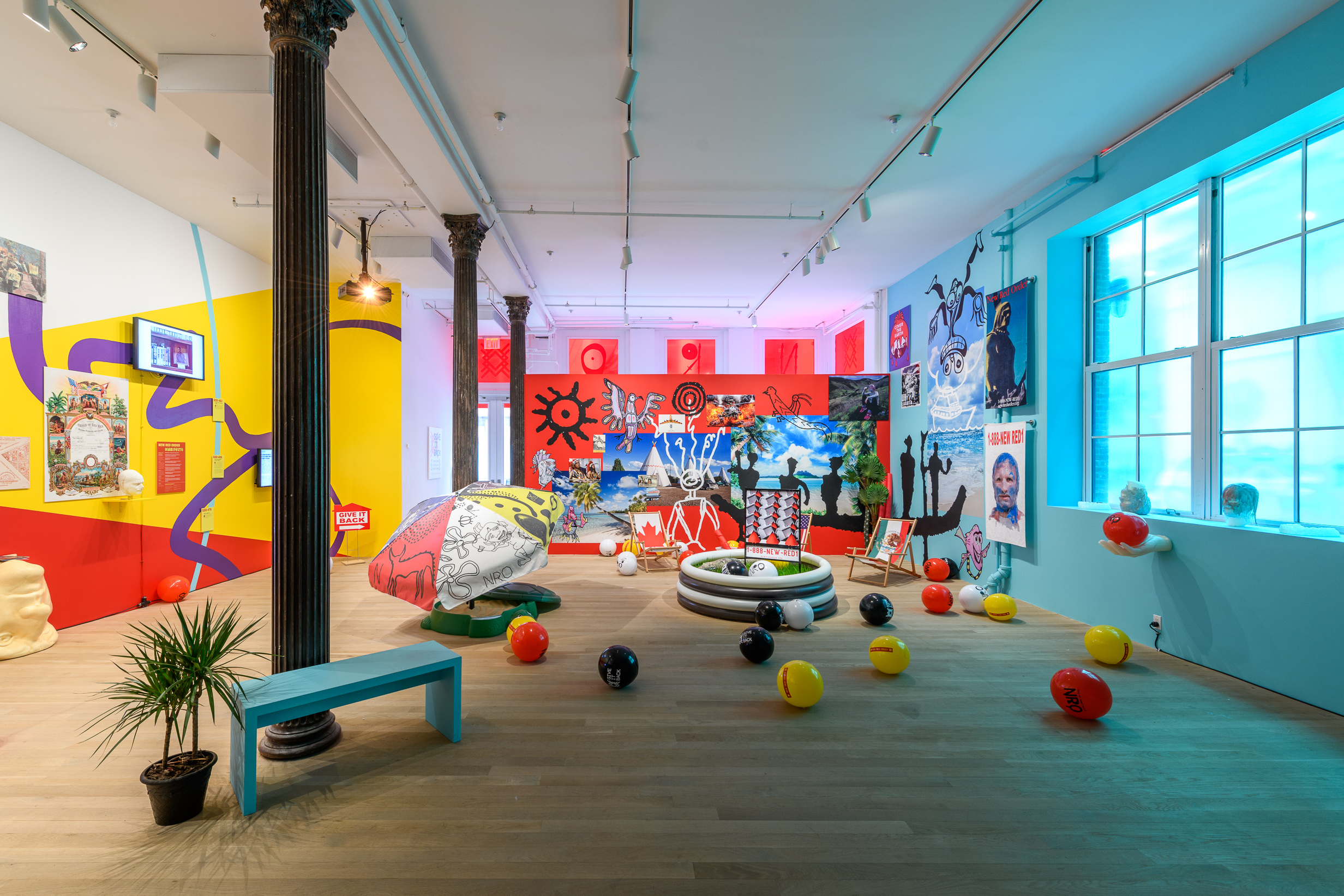 A wide angle image of a multicolored exhibition space consisting of scattered beach balls, an inflatable pool, a beach umbrella, a beach chair, numerous wall graphics and tv monitors.