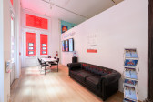 A wide angle image of a mockup real estate office consisiting of a leather couch, a magazine rack, a table and multiple tv monitors. The windows are lined with real estate listings.