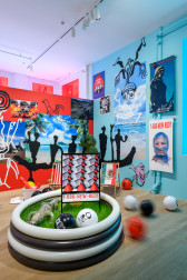 An image of a multicolored exhibition space consisting of beach paraphernalia, a real estate lawn sign, and a mural wall collaged with images and drawings