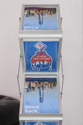 An image of a magazine rack with numerous copies of a magazine with a city-scape is positioned upside down, the point of the skyscraper reaching towards the bottom of the image. Googley eyes, eyebrows, and a mouth are drawn near the top of the building. Red serif font across the top of the image reads 