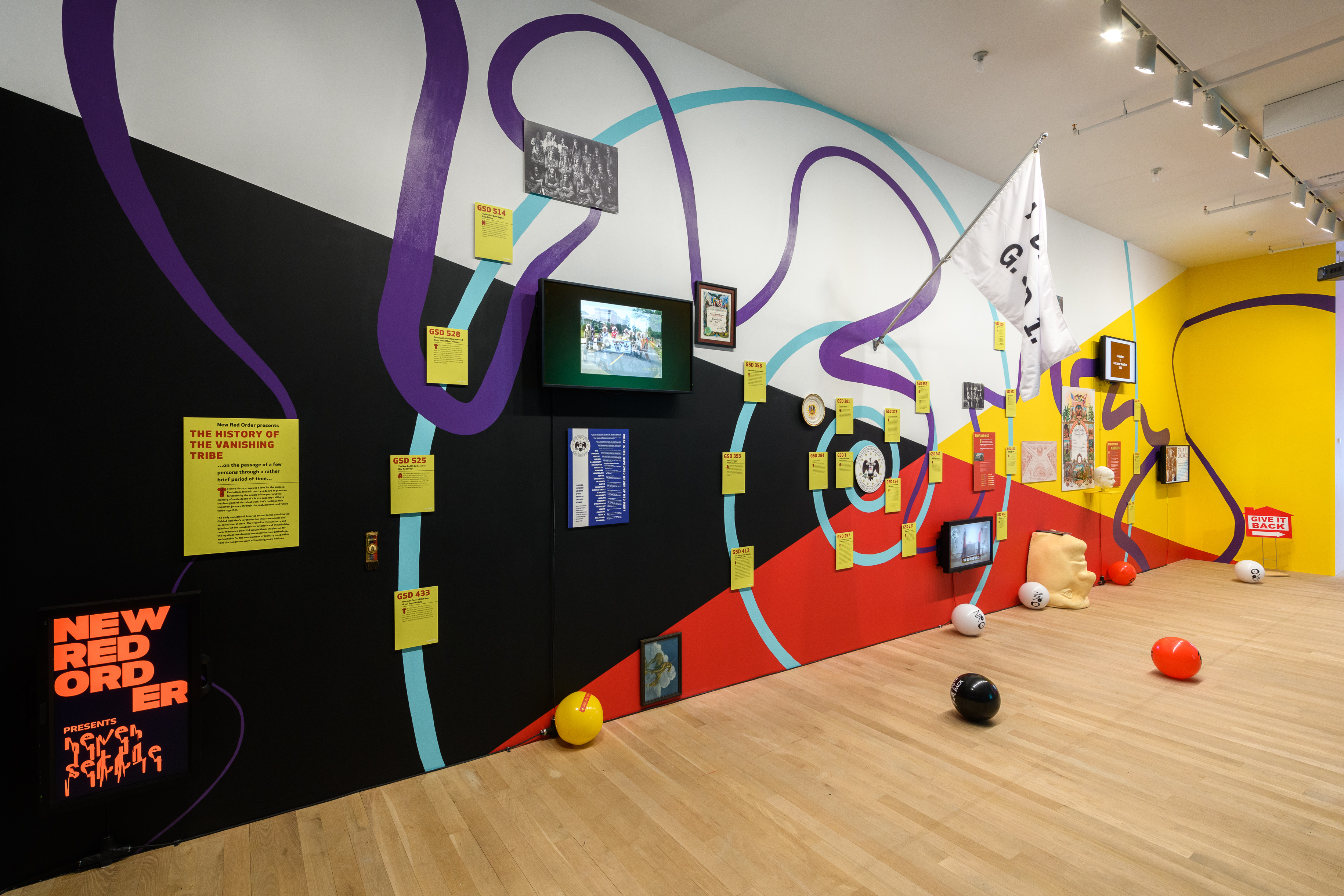 An image of a multicolored wall filled with multiple tv monitors and info graphics. Black, red and yellow beach balls are scattered along the floor.