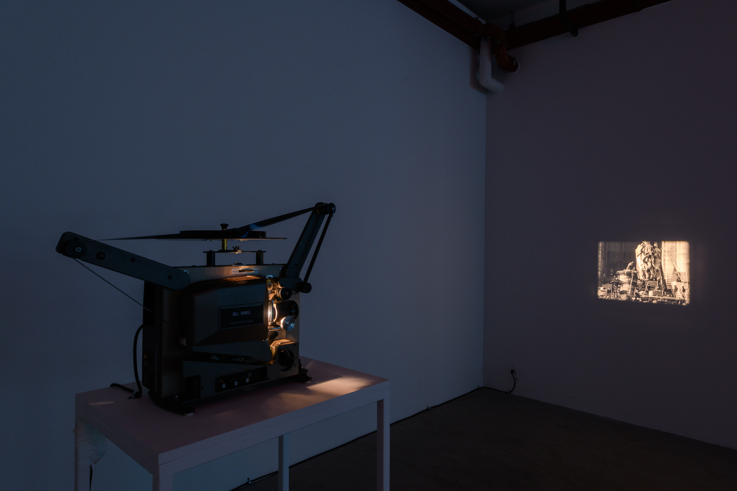 An image of a dark exhibition space, a film projector projects a sepia tone image onto the wall.