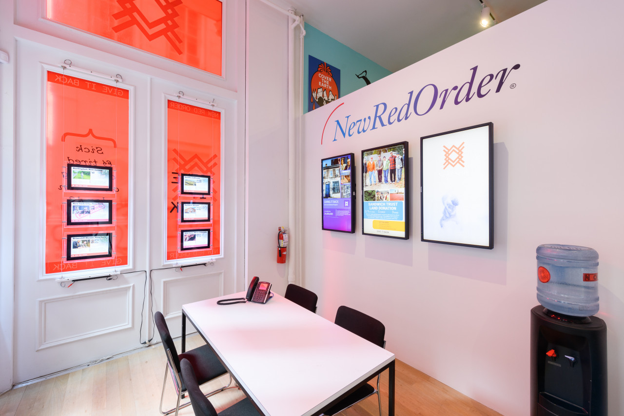 A wide angle image of a mockup real estate office consisiting of a table,chairs, a water coolor, and multiple tv monitors. The windows are lined with real estate listings and on the wall reads "New Red Order"..
