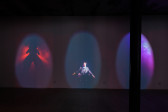 An image of a dark exhibition space with three video projections projected on to a wall.