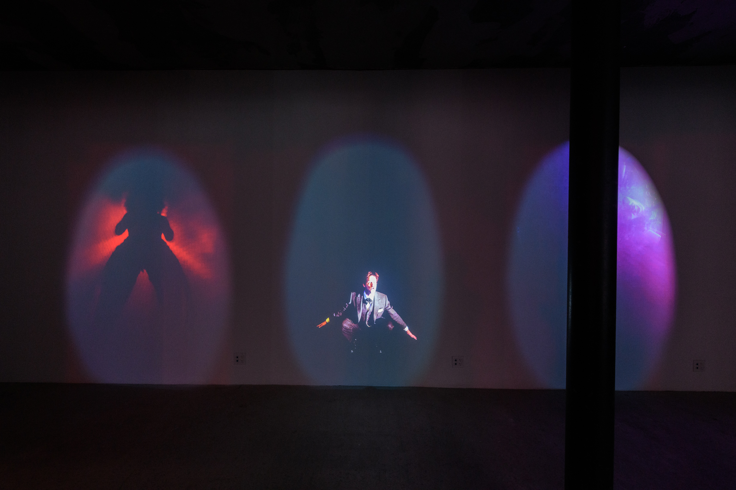 An image of a dark exhibition space with three video projections projected on to a wall.