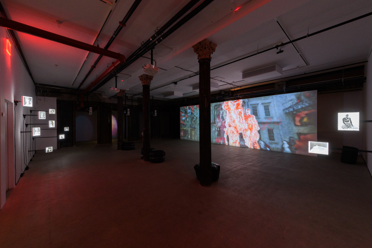 A wide angle image of a dark exhibition space, on the left a large projection is shown on a long wall, on the right photograph negatives hang off a wall.