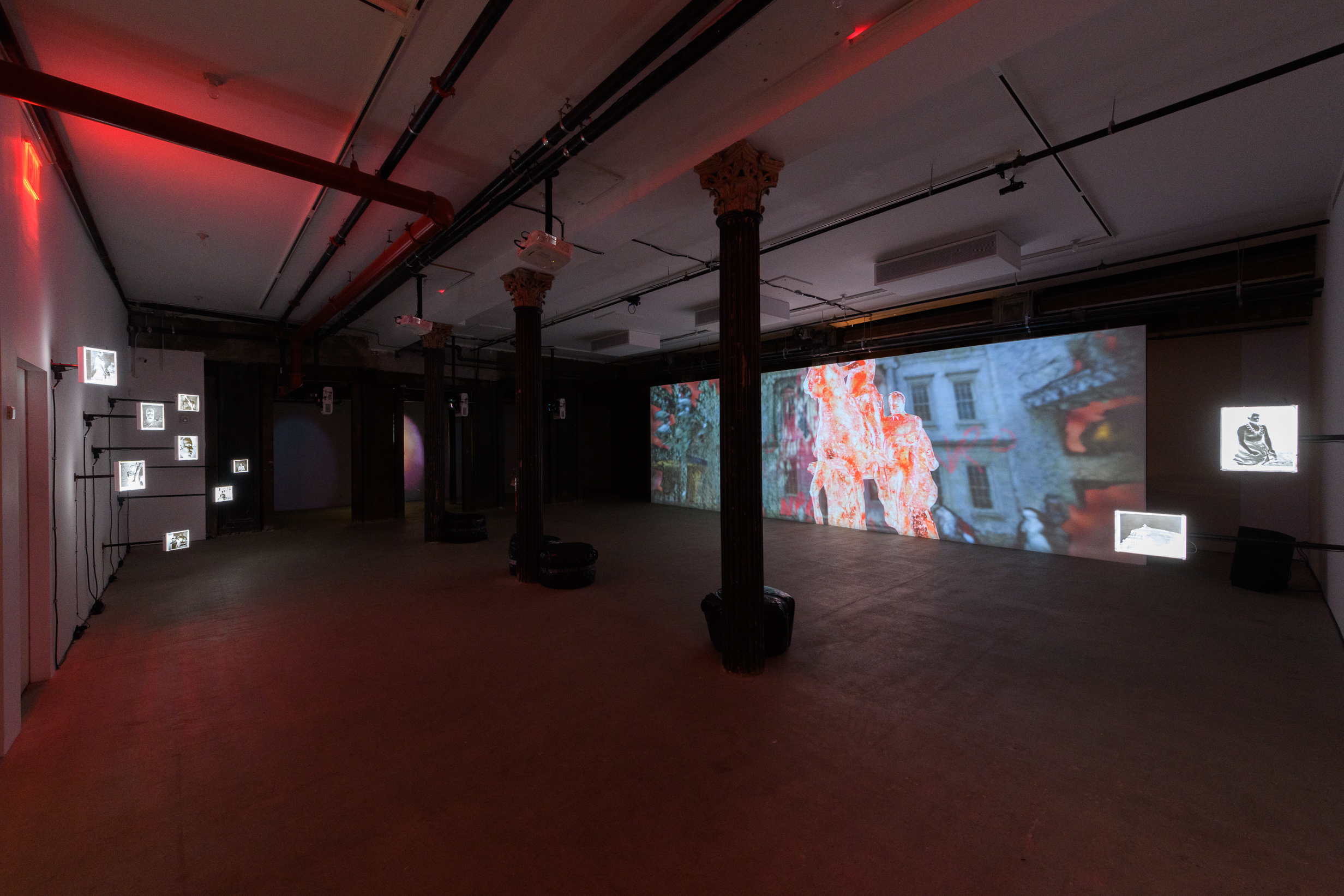 A wide angle image of a dark exhibition space, on the left a large projection is shown on a long wall, on the right photograph negatives hang off a wall.
