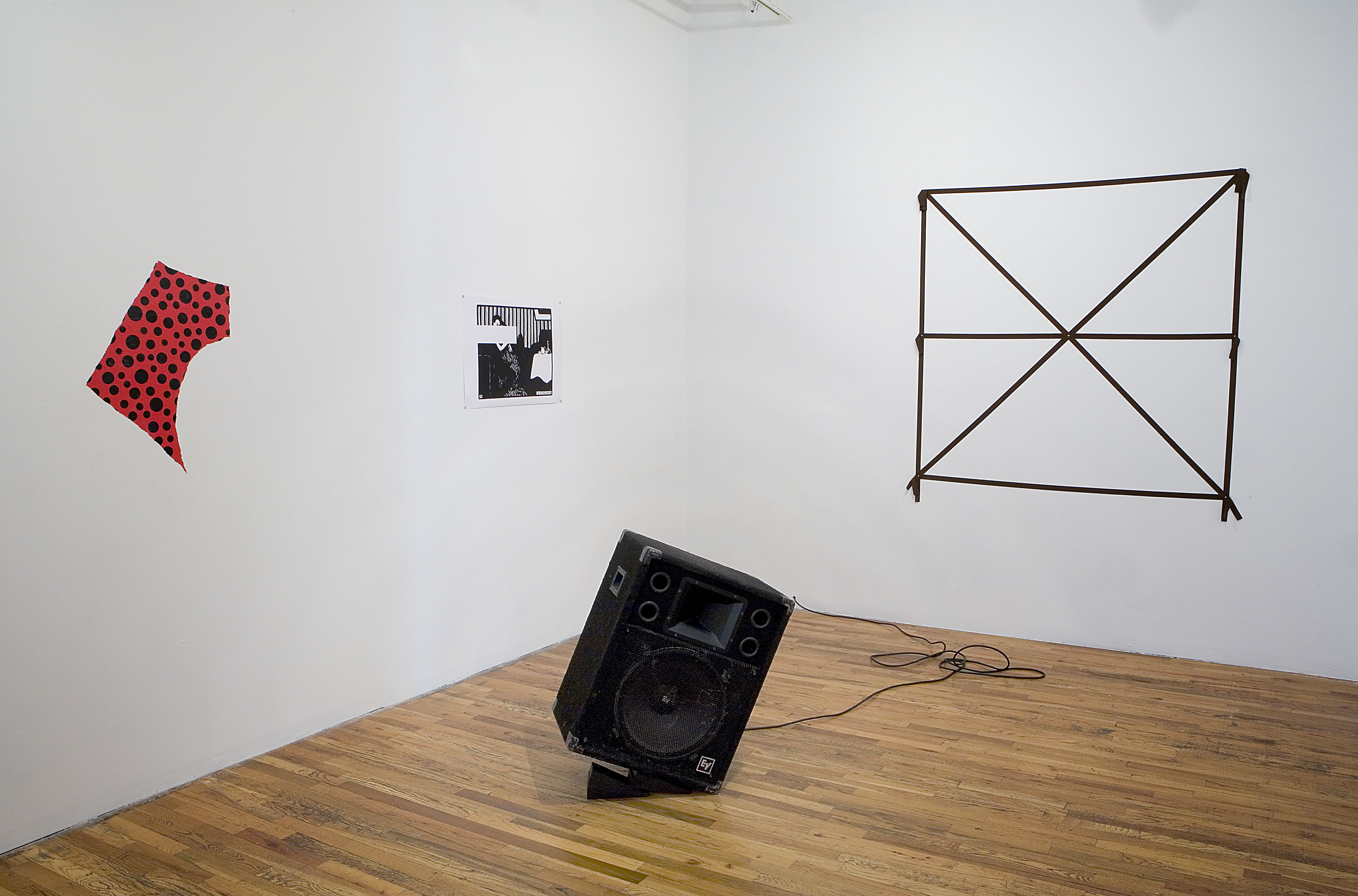 A large black speaker rests at an angle in the middle of a wood floor. On the walls, a red cloth with black polka dots, a black and white photograph, and a taped black square with an X and horizontal line through the center are displayed.