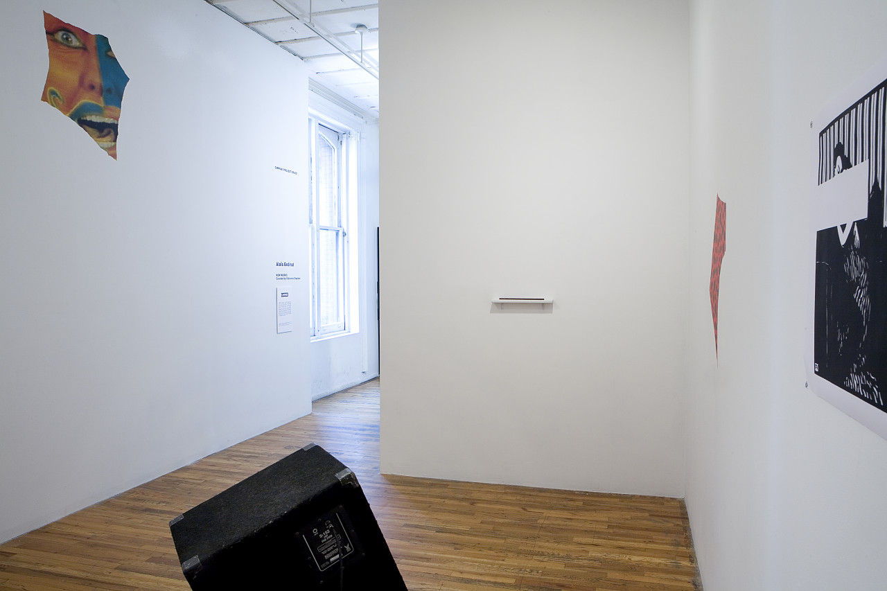 A black cube-shaped speaker sits, tilted upwards, in the middle of a room with wooden floors and white walls. On a wall to the left of the image is a printed image of a fragment of a face with an open mouth, the skin painted in red, blue, and yellow. On a wall to the right are two printed works, one an illustration of a figure in black and white but with a section of the image above the figure's head blocked out with white space, another is a red and blue polka dot pattern in a hand-cut shape. On the wall in the middle of the room is a small white shelf with a small flat object on top of it.
