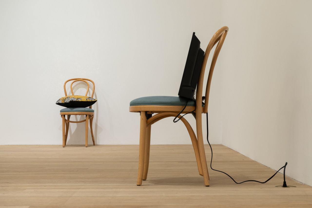 A color photograph of two wooden cafe chairs with green cushions in a gallery space. A television screen with a cord extending down to an outlet in the floor rests upon the chair closest to the camera. A pillow with black backing and a gold-silver sequined front rests on the chair in the background.