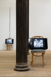 A color photograph of two wooden cafe chairs with green cushions in a gallery space with a dark metal column. A television screen with a cord extending down to an outlet in the floor rests upon each chair.