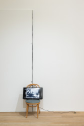 A color photograph of a wooden cafe chair with a green cushion in a gallery space. The chair is placed in front of a small seam in the wall. A television screen rests on top of the chair.