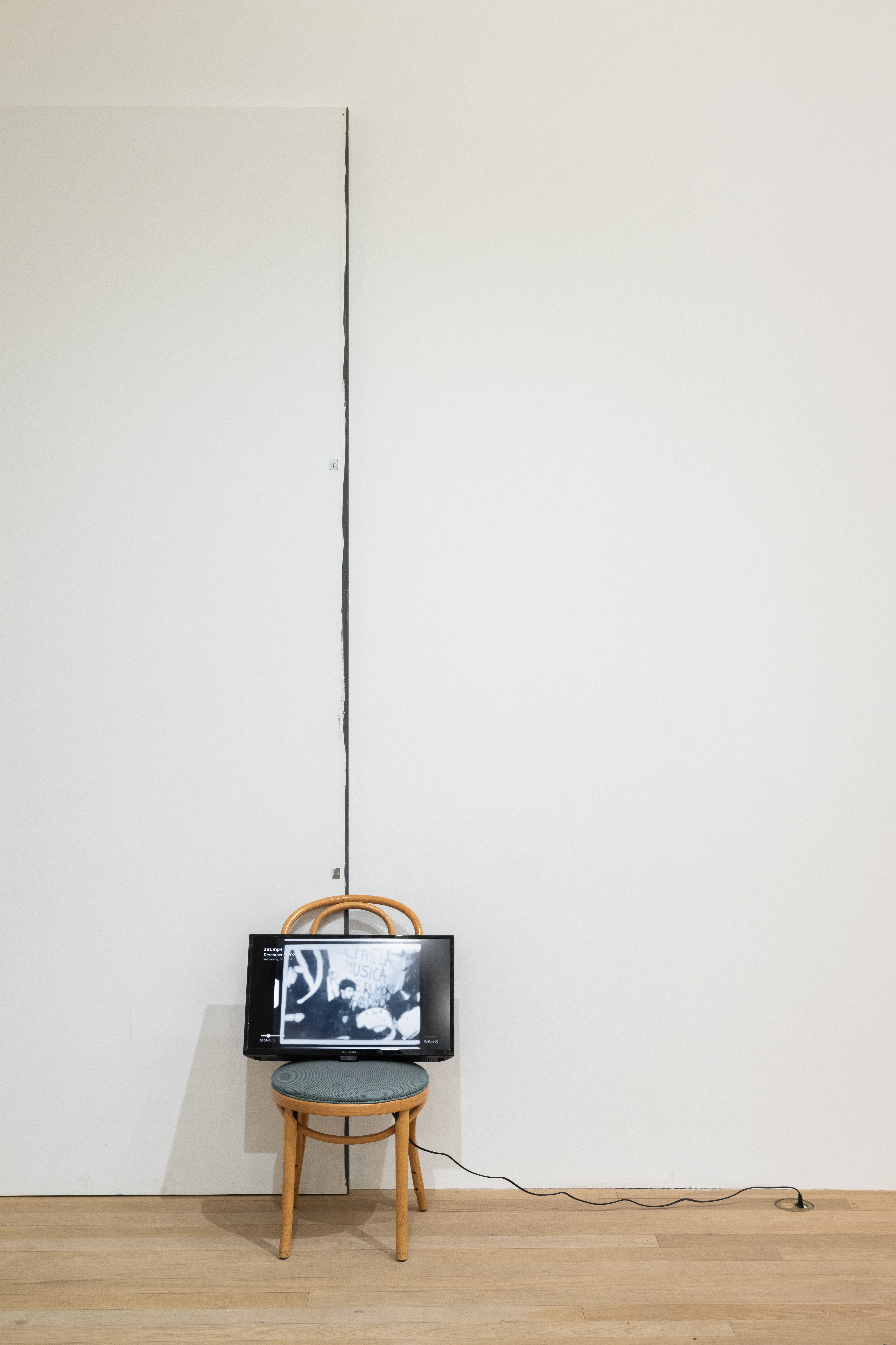 A color photograph of a wooden cafe chair with a green cushion in a gallery space. The chair is placed in front of a small seam in the wall. A television screen rests on top of the chair.
