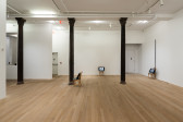 A color photograph of three chairs spread out in a gallery space with three dark columns. A television screen rests on each of the chairs.