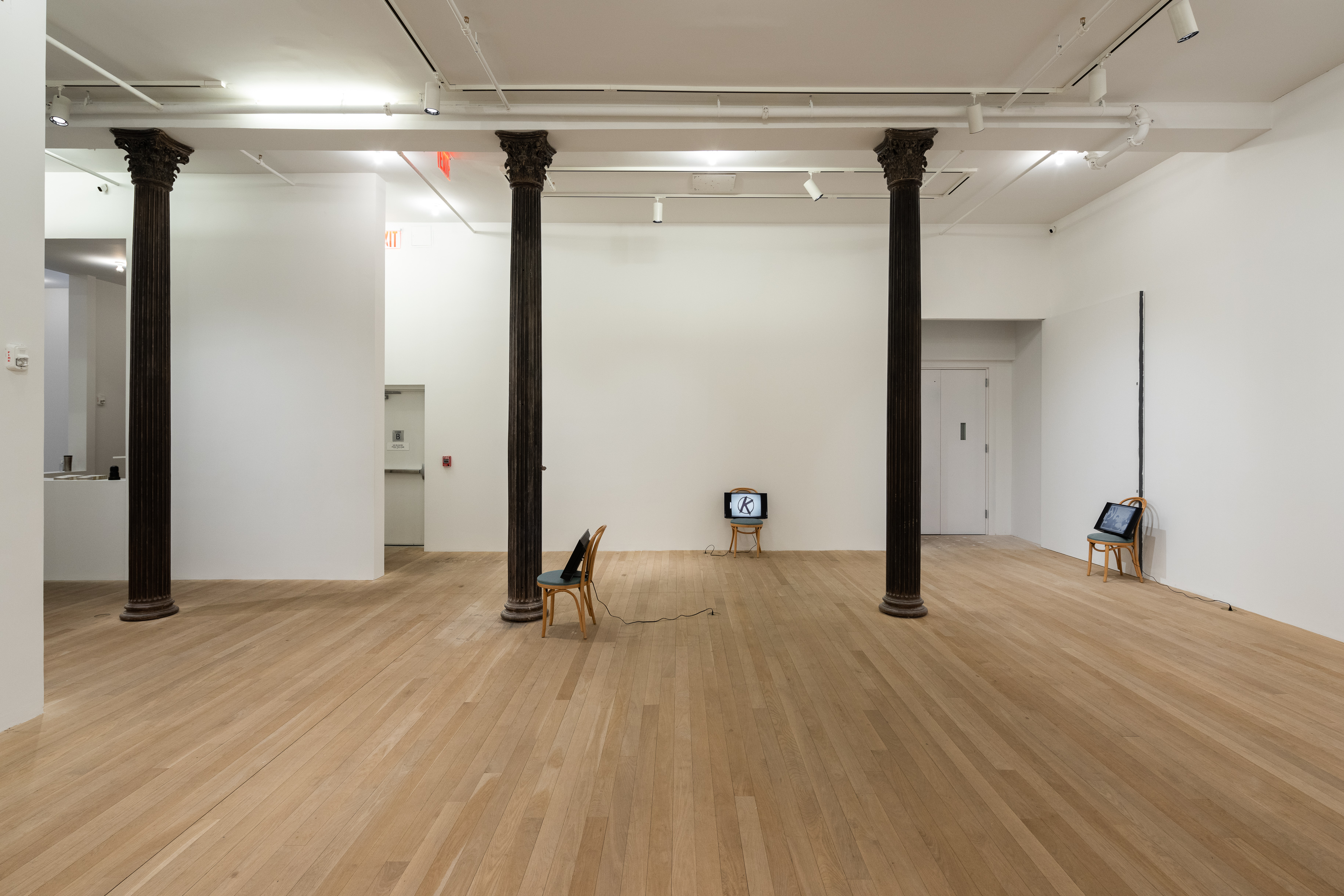 A color photograph of three chairs spread out in a gallery space with three dark columns. A television screen rests on each of the chairs.