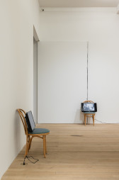 A color photograph of two wooden cafe chairs with green cushions in a gallery space. A television screen with a cord extending down to an outlet in the floor rests upon each chair.