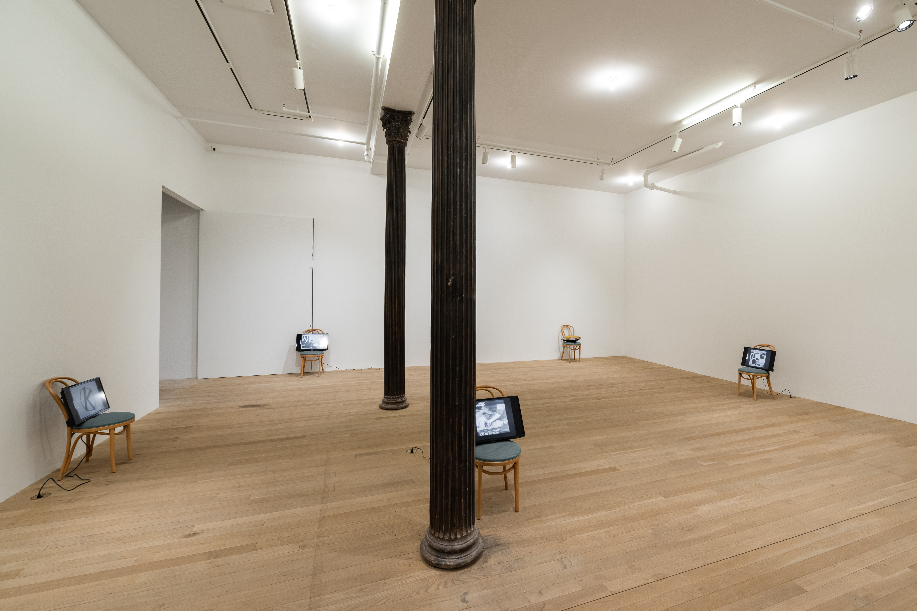 A color installation photograph of five chairs spread out in a gallery space bisected by two dark columns. The chairs have tv screens resting on their green cushions.