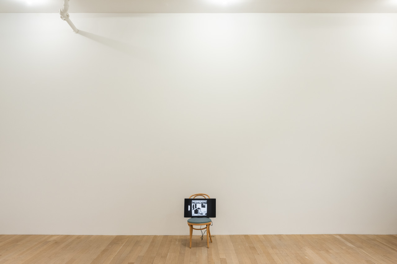 A color photograph of a wooden cafe chair with a green cushion in a gallery space. A television screen rests on top of the chair.