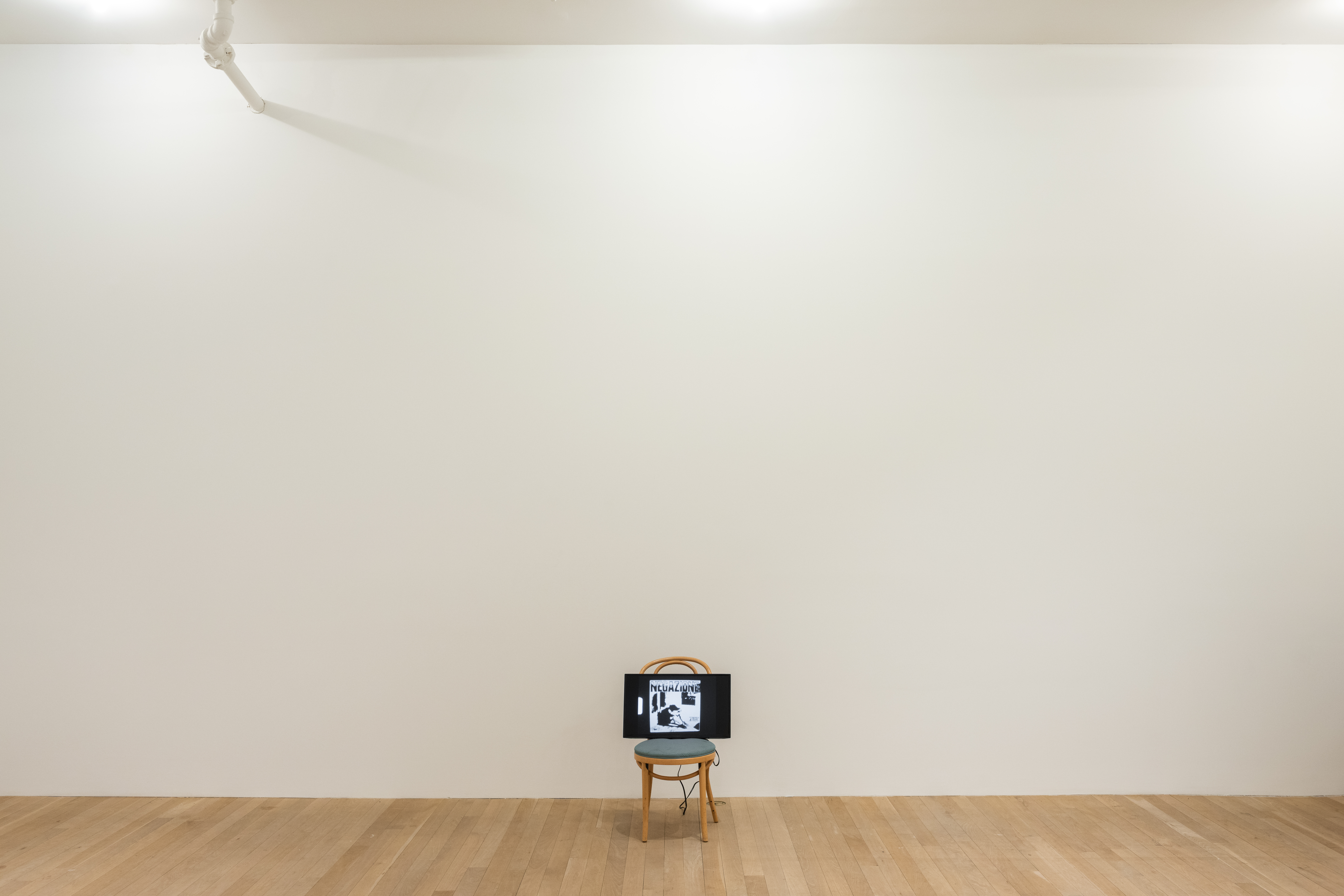 A color photograph of a wooden cafe chair with a green cushion in a gallery space. A television screen rests on top of the chair.