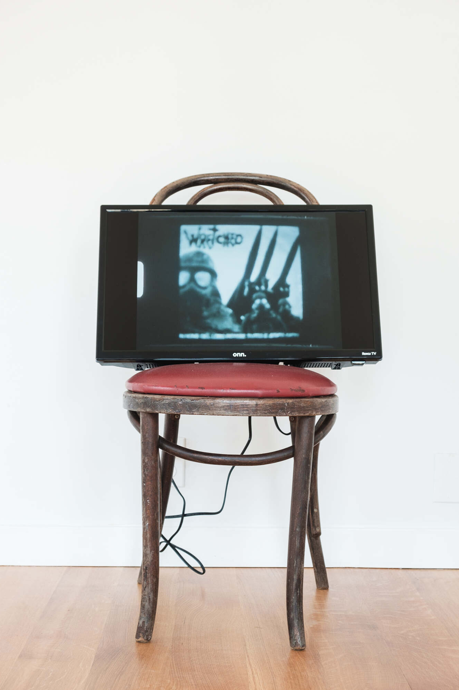 A color photograph of a television screen resting on a wooden chair with a red cushion. The screen displays a black and white image of several masked figures.