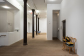 A color photograph of a gallery space with a row of dark columns on the left side. Several wooden cafe chairs with green cushions are visible. Upon the chairs rest gold-silver sequined pillows or television screens.