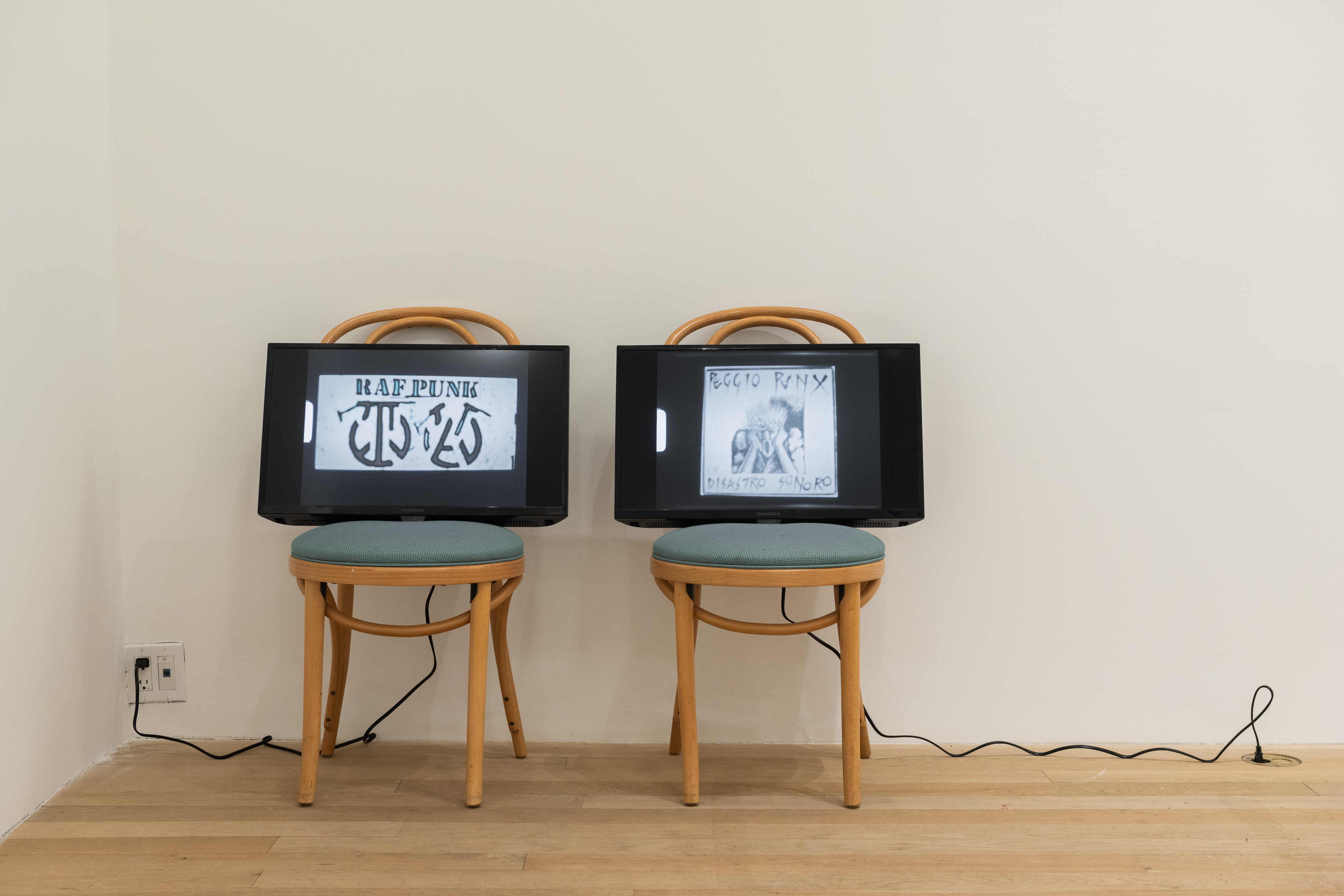 A color photograph of two wooden cafe chairs with green cushions in a gallery space. A television screen with a cord extending down to an outlet in the floor rests upon each chair.