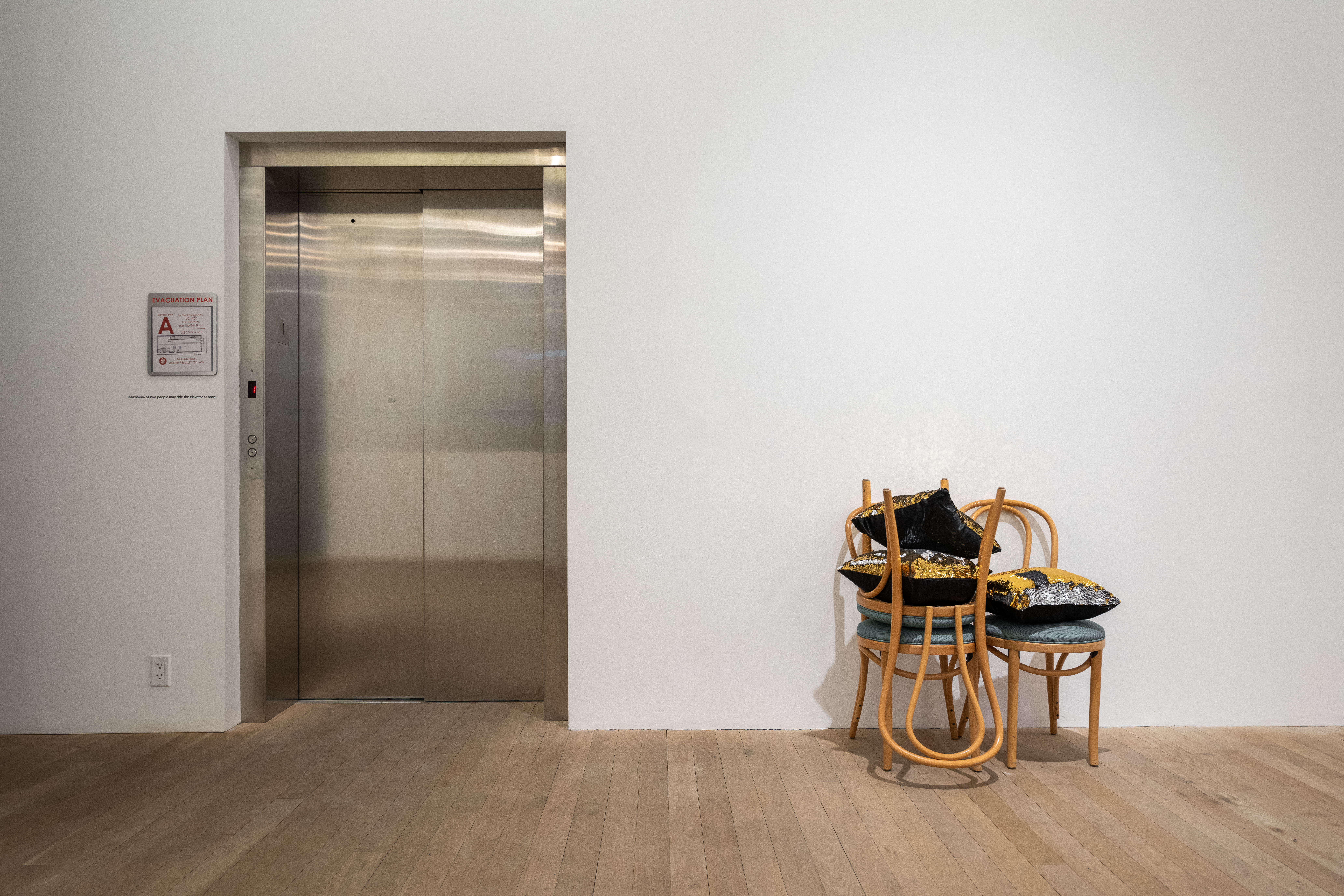 A color photograph of three wooden cafe chairs with green cushions next to a metal elevator door. One chair is stacked upside down on another on the left, and two pillows with black backs and sequined gold-silver fronts are placed between the chair's upturned legs. Another pillow rests on the cushion of the chair on the right.