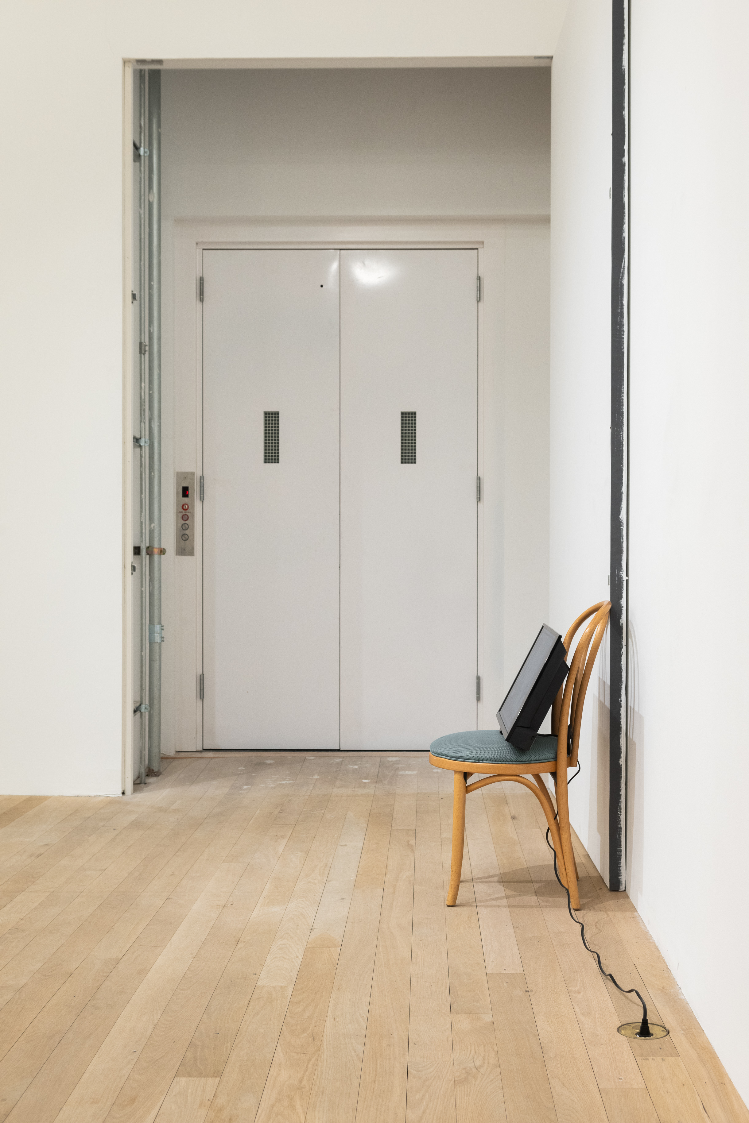 A color photograph of a wooden cafe chair with green cushion in a gallery space. The chair is placed up against the wall and a set of doors is visible in the background. A television screen rests atop the chair with a cord extending down to an outlet on the ground.