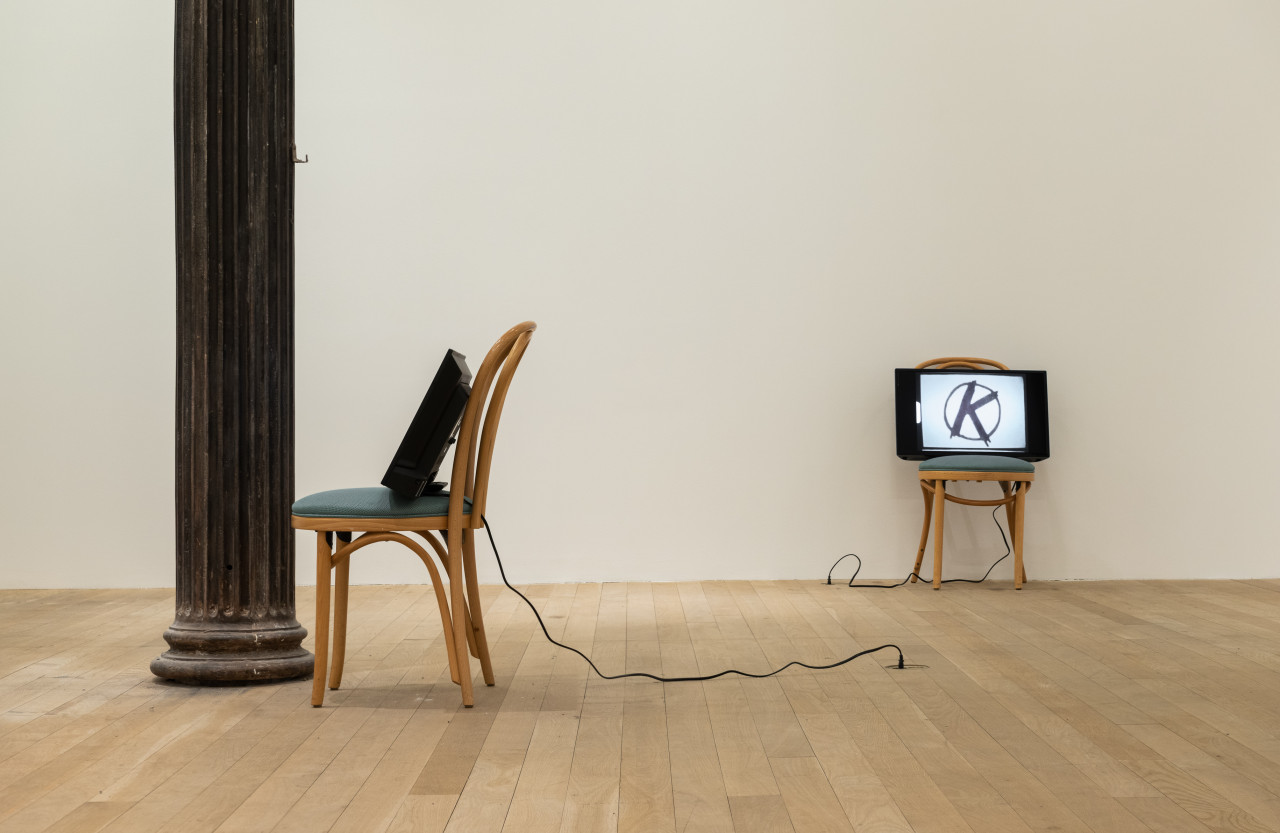 A color installation photograph of two wooden cafe chairs with green cushions in a gallery space with a column on the left. Resting on the chairs are two television monitors with cords that extend to outlets on the ground.