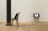 A color installation photograph of two wooden cafe chairs with green cushions in a gallery space with a column on the left. Resting on the chairs are two television monitors with cords that extend to outlets on the ground.