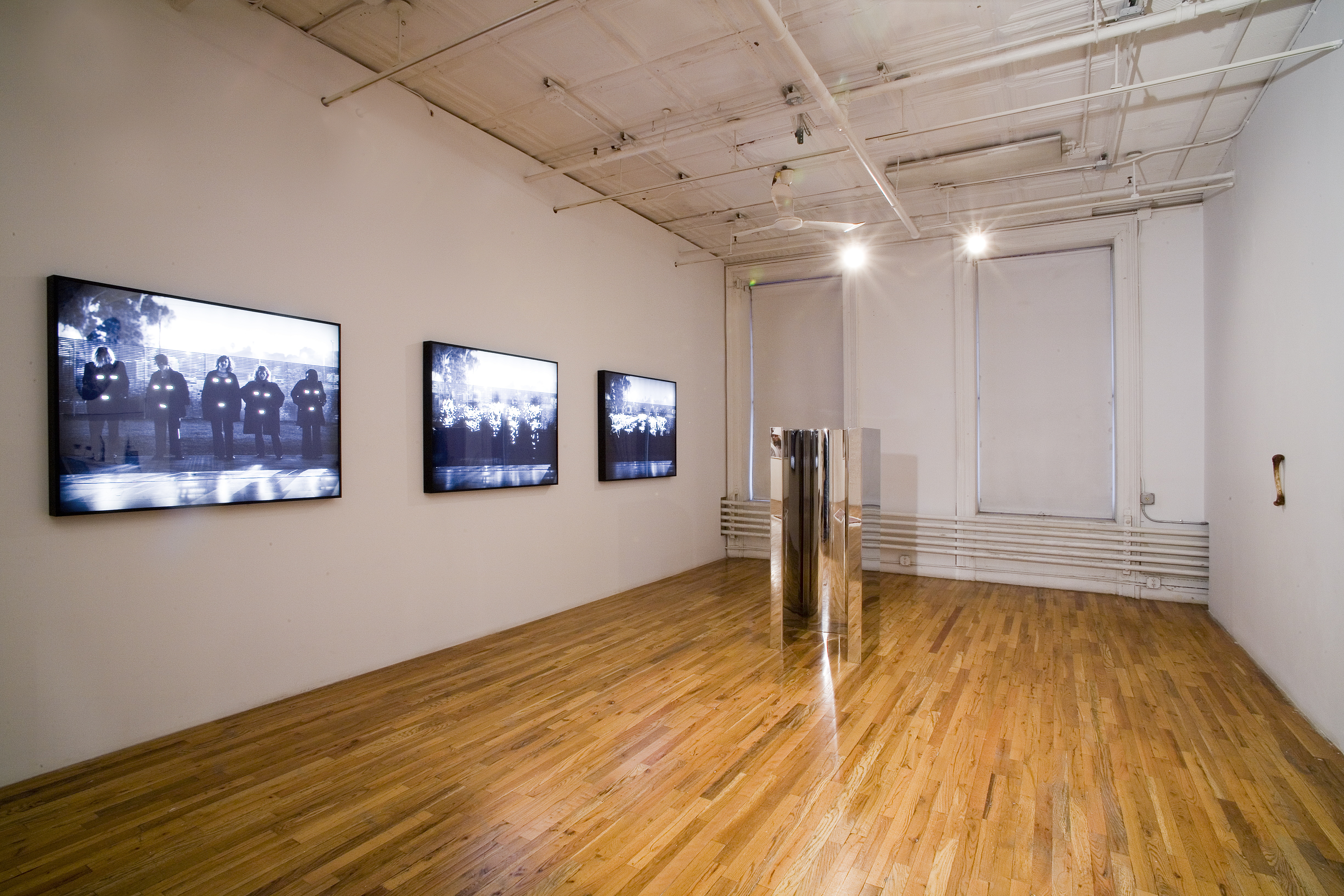 Three screens and a metallic sculpture are installed in a gallery space with wooden floors. The three screens are arranged next to each other, with the screen closest to the viewer displaying a series of figures with three bright forms attached to their torsos. The other two screens show increasingly blurry renderings of the same image. To the right of the screens, a tall, shiny and metallic sculpture sits on the gallery floor. A pocket-sized image of a figure is pasted to the sculpture's left sides.