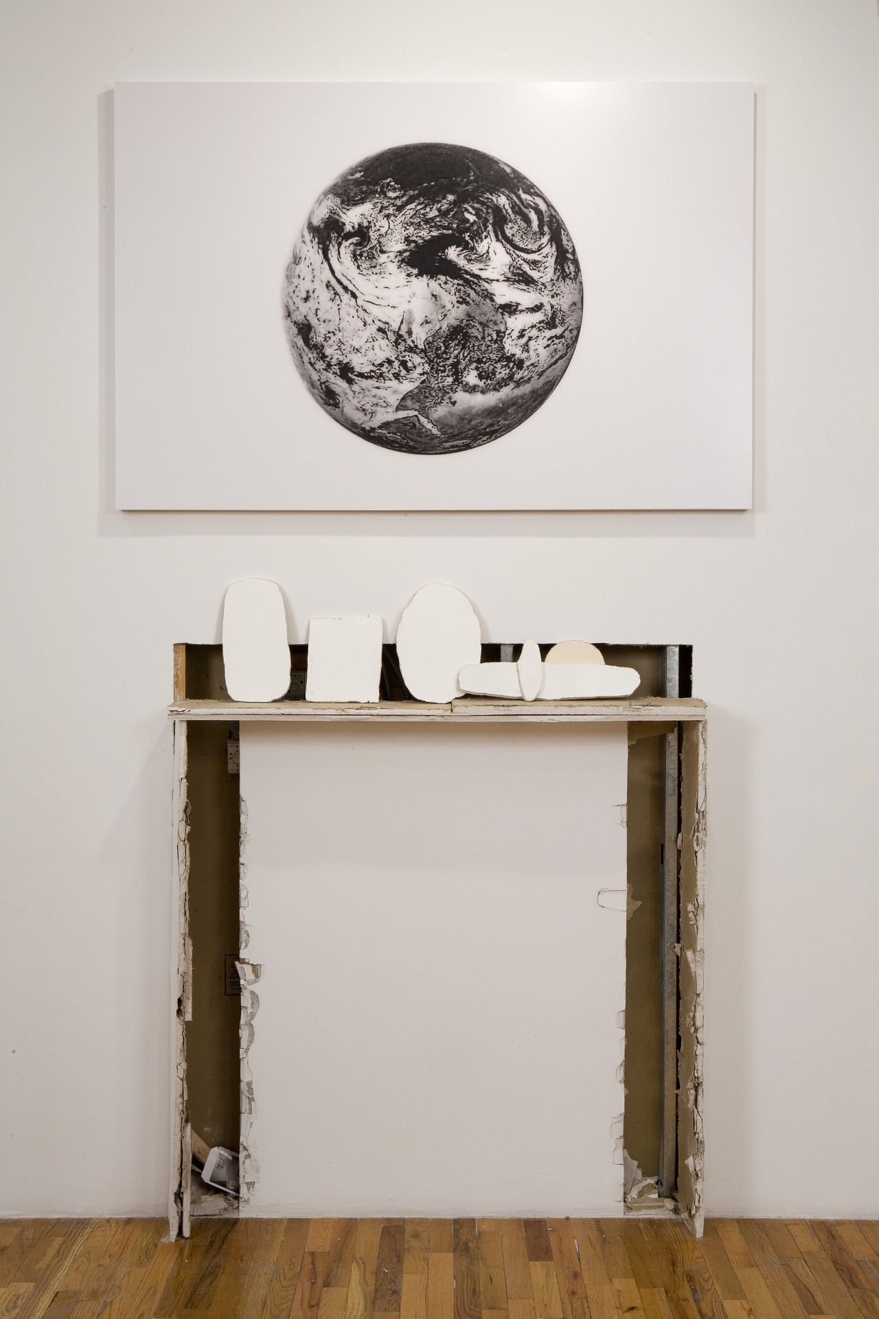 Close up of an artwork that consists of a white, rectangular canvas with a black and white image of a planet with a swirling, cloudy atmosphere. Directly below, a shelf made out of worn wooden planks supports several rough, oval and square shaped 2D cutouts that are arranged next to one another. A strip of wall behind the shelf and two "leg" like shapes below it have been gouged out of the gallery wall, revealing insulation and building supports.
