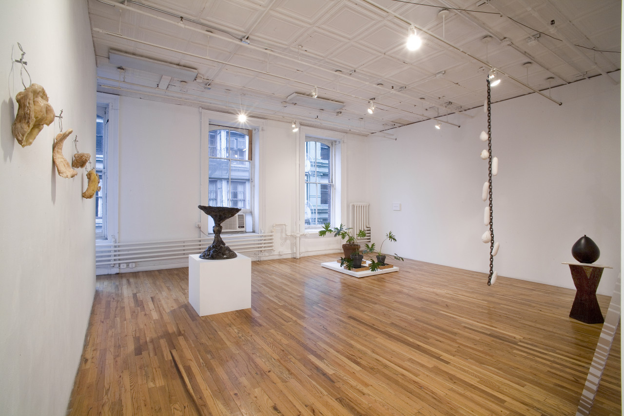 A series of sculptures in a large gallery space with wooden floors. In the center of the image, a dark sculpture forming an hourglass shape rests on a white plinth, while three potted ferns rest on another, low plinth to the right. A long chain with white appendages hangs from the gallery ceiling.