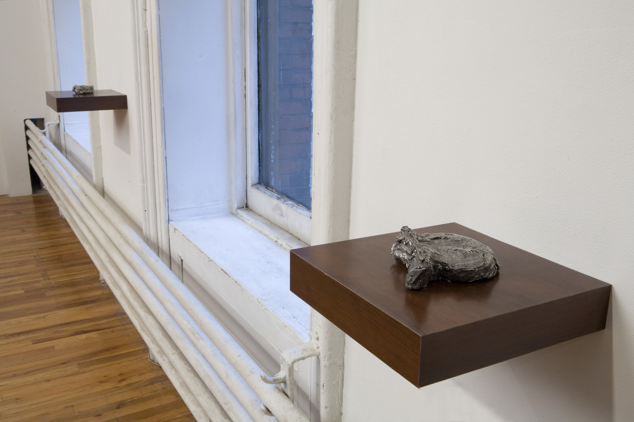 Close up of a small sculpture resting on a square wooden support jutting out from a white gallery wall next to a window. The sculpture has a metallic color and an irregular, round shape, with a tree-bark like texture. A similar sculpture and support are mounted on the other side of the gallery window.