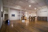 Two shelves and several artworks sit in a gallery space with wooden floors. To the right, there are two empty wooden bookshelves placed at right angles to one another. Above a bookshelf positioned against the gallery wall, there is text that reads 