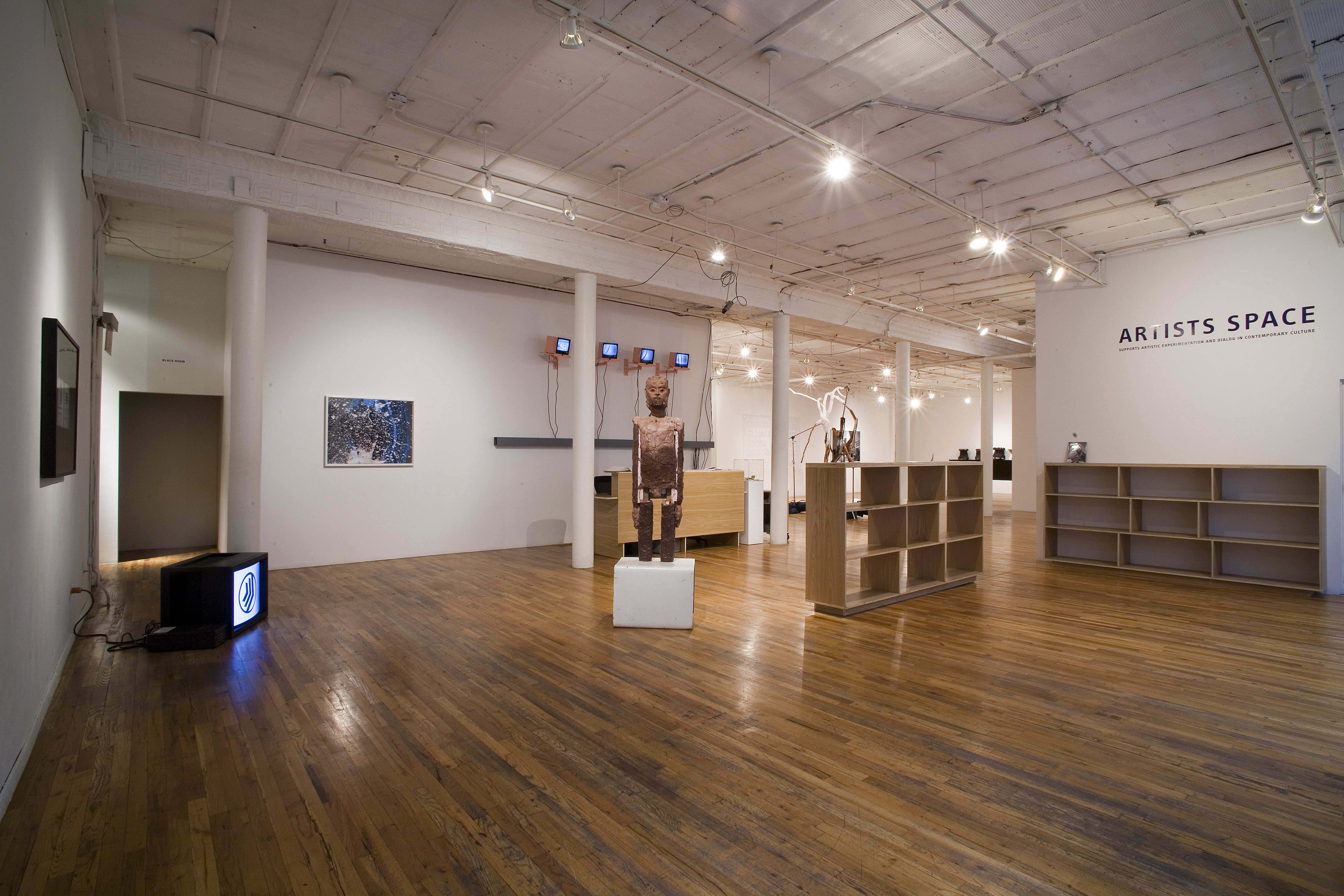 Two shelves and several artworks sit in a gallery space with wooden floors. To the right, there are two empty wooden bookshelves placed at right angles to one another. Above a bookshelf positioned against the gallery wall, there is text that reads 