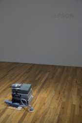 On a wooden floor in a white room are a pile of objects which emulate video projectors plugged into a power strip nearby. There is a remote control on the pile. On the otherwise empty facing white wall, 