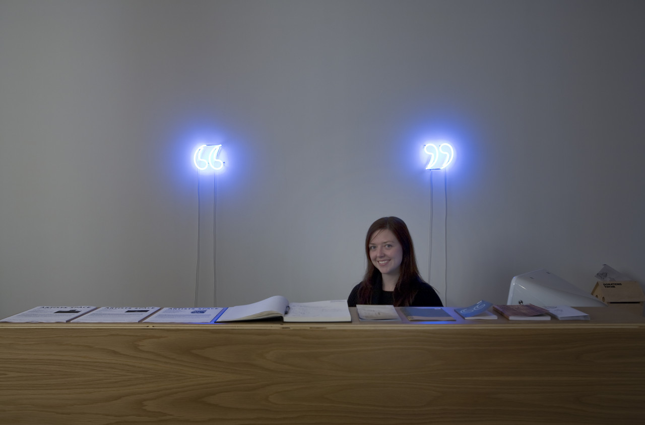 A wooden reception desk with piles of various materials on it. A person sits at the desk smiling, and behind them, mounted on the wall, are two glowing neon quotation marks.