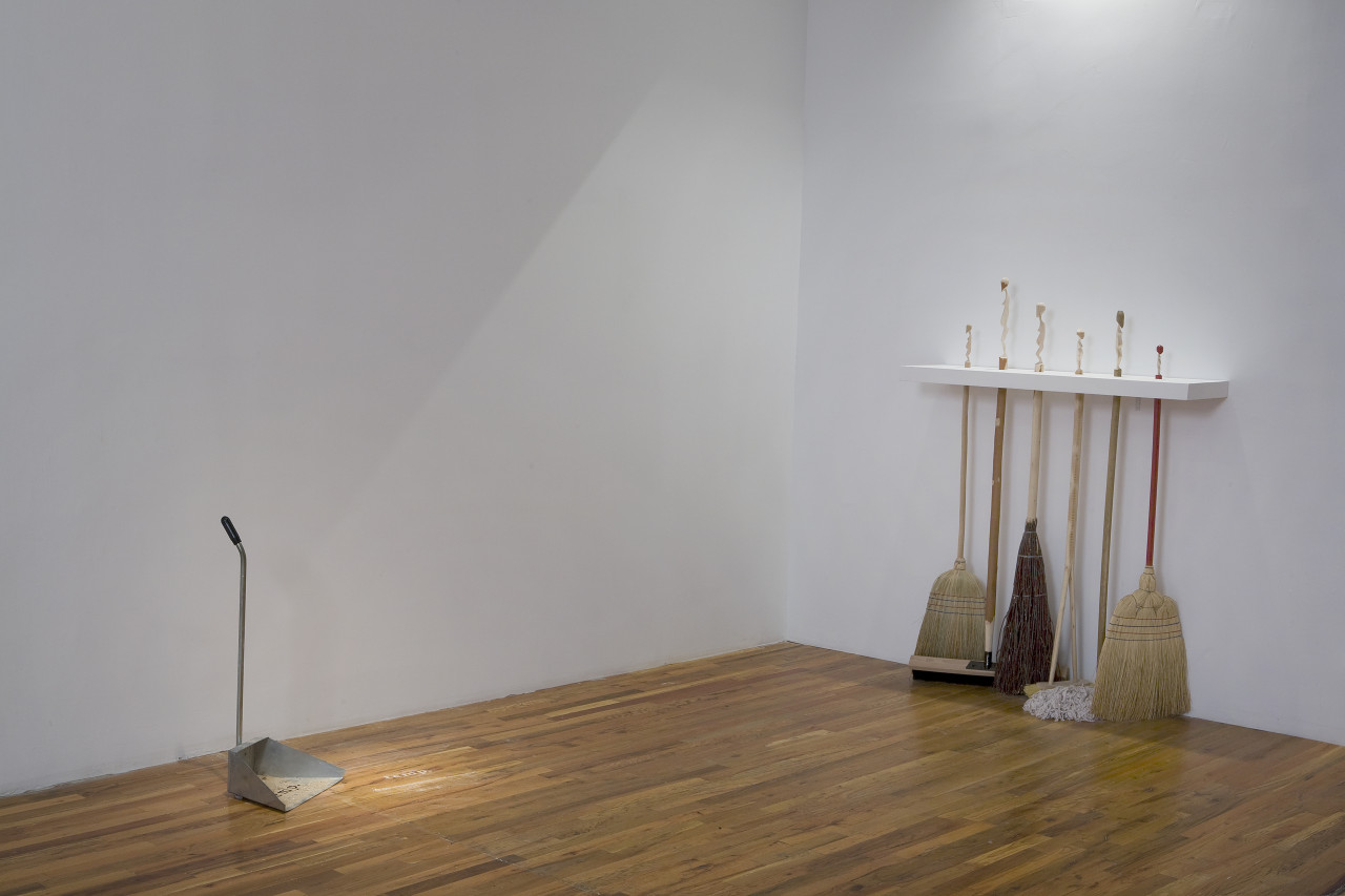 In the corner of a room, a group of six brooms and mops is held free-standing off of a wall by a shelf which allows the handles to pass through it, holding them in place in a line. Near the adjacent wall, a metal dustbin stands facing the brooms, dust both within the dustbin and nearby.