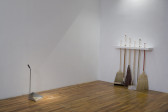 In the corner of a room, a group of six brooms and mops is held free-standing off of a wall by a shelf which allows the handles to pass through it, holding them in place in a line. Near the adjacent wall, a metal dustbin stands facing the brooms, dust both within the dustbin and nearby.