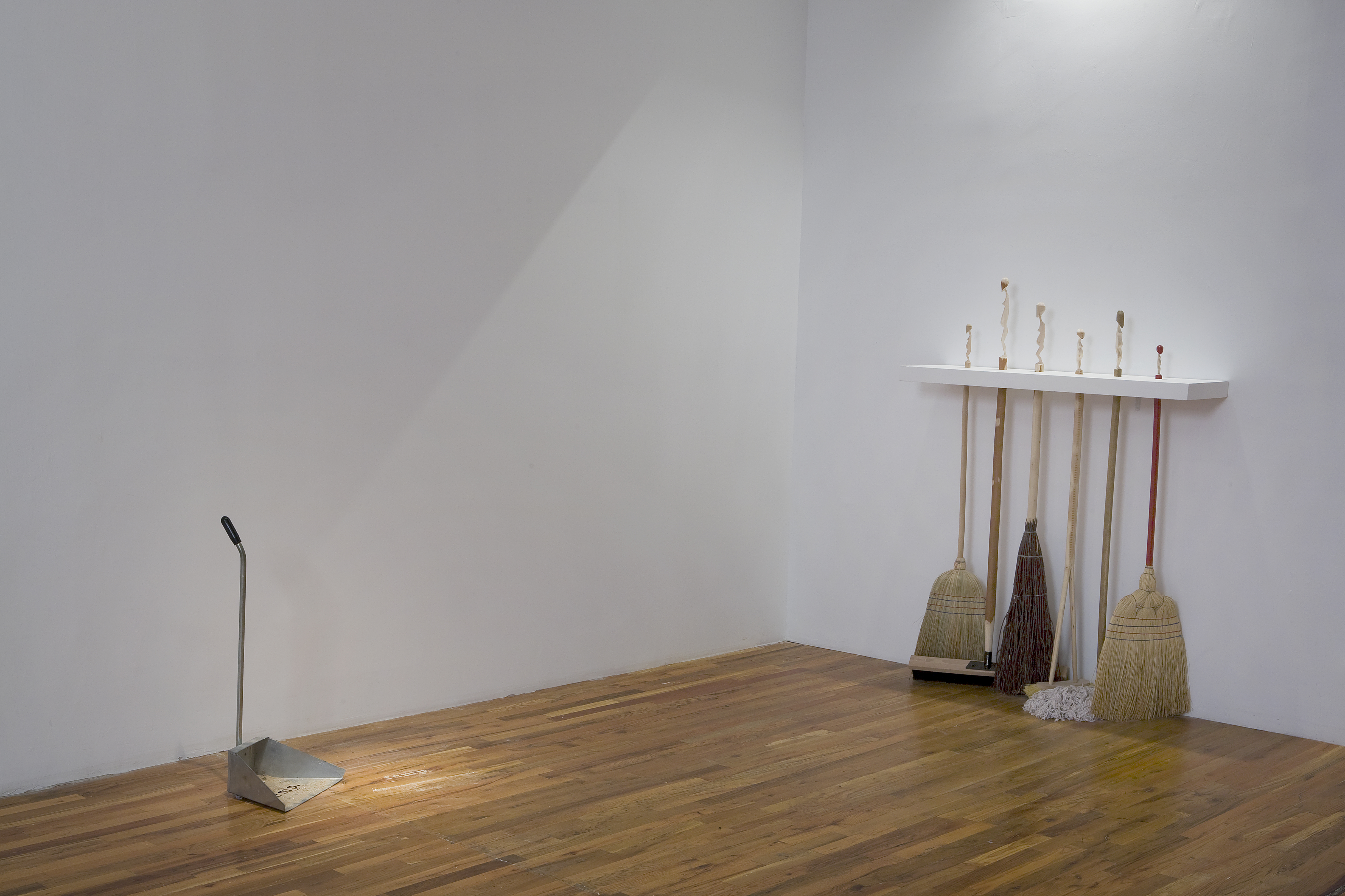 In the corner of a room, a group of six brooms and mops is held free-standing off of a wall by a shelf which allows the handles to pass through it, holding them in place in a line. Near the adjacent wall, a metal dustbin stands facing the brooms, dust both within the dustbin and nearby.