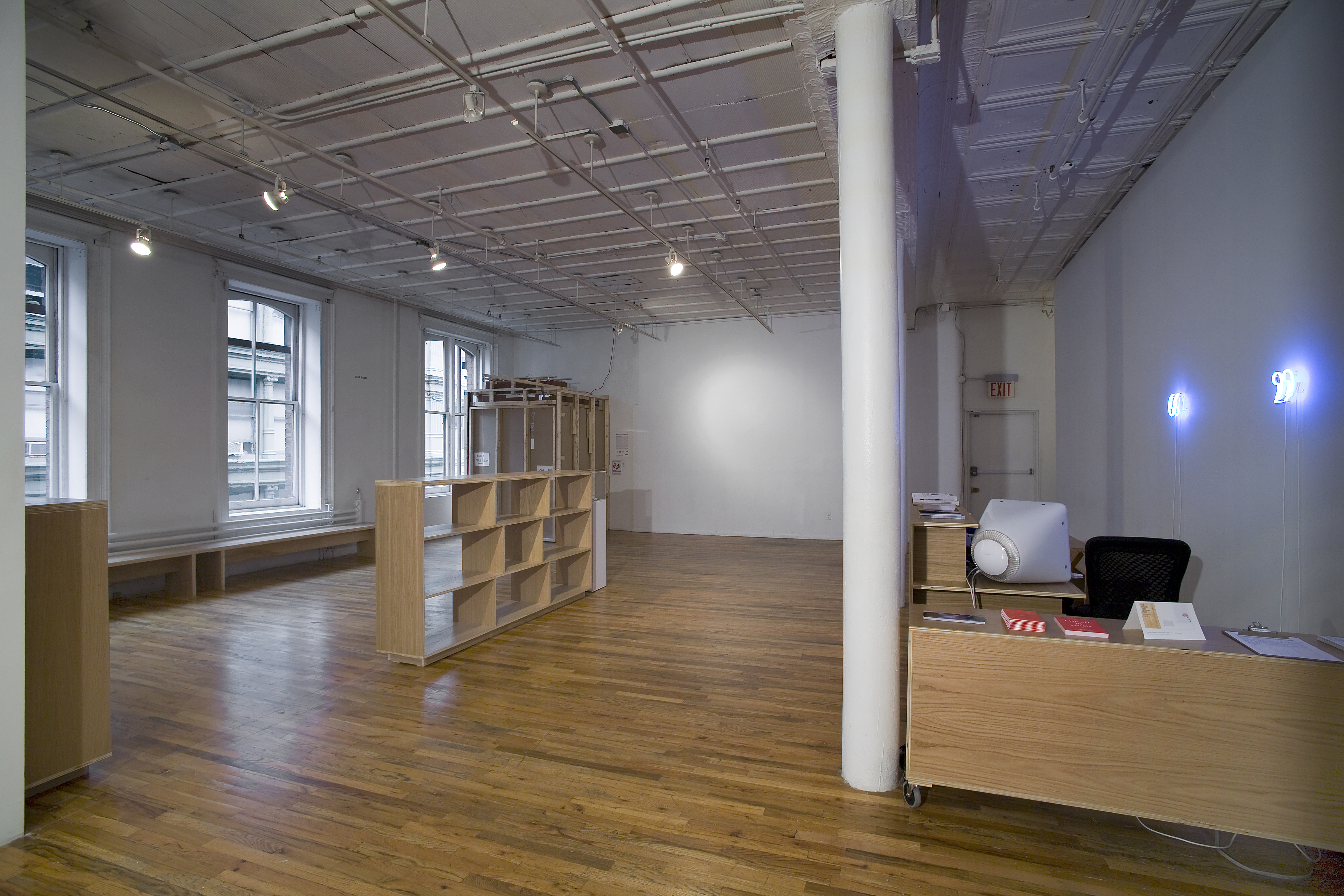 The interior of a Soho loft space with a wooden front desk to the right. Two neon quotation marks hang behind it. Within the room there are empty wooden shelves, wooden benches, and a rough-hewn wooden enclosure at the far end.