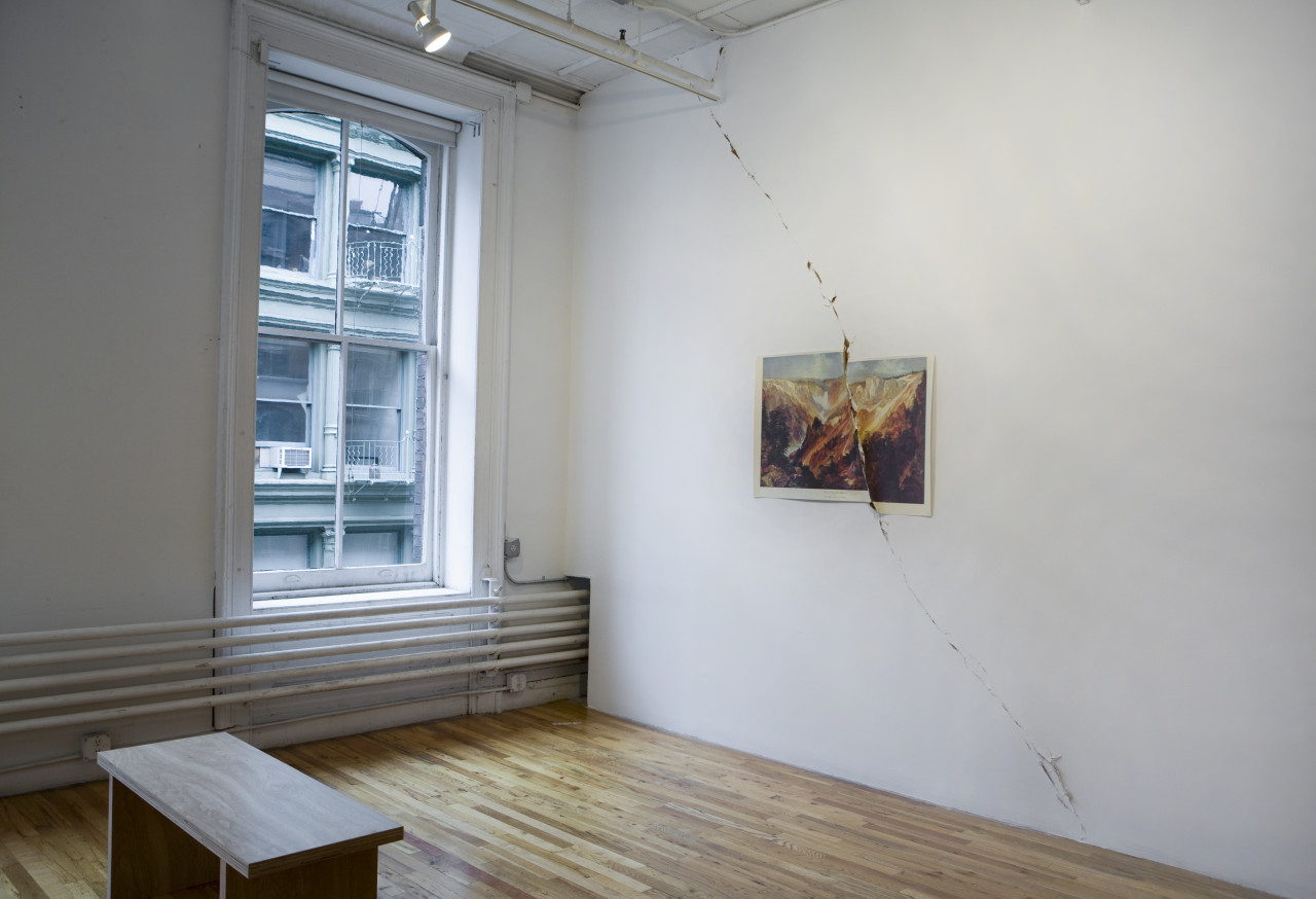 The corner of a loft space. A printed image of a canyon is affixed to the wall. The image is torn in half and the tear extends off of the paper onto the wall itself, continuing from floor to ceiling.