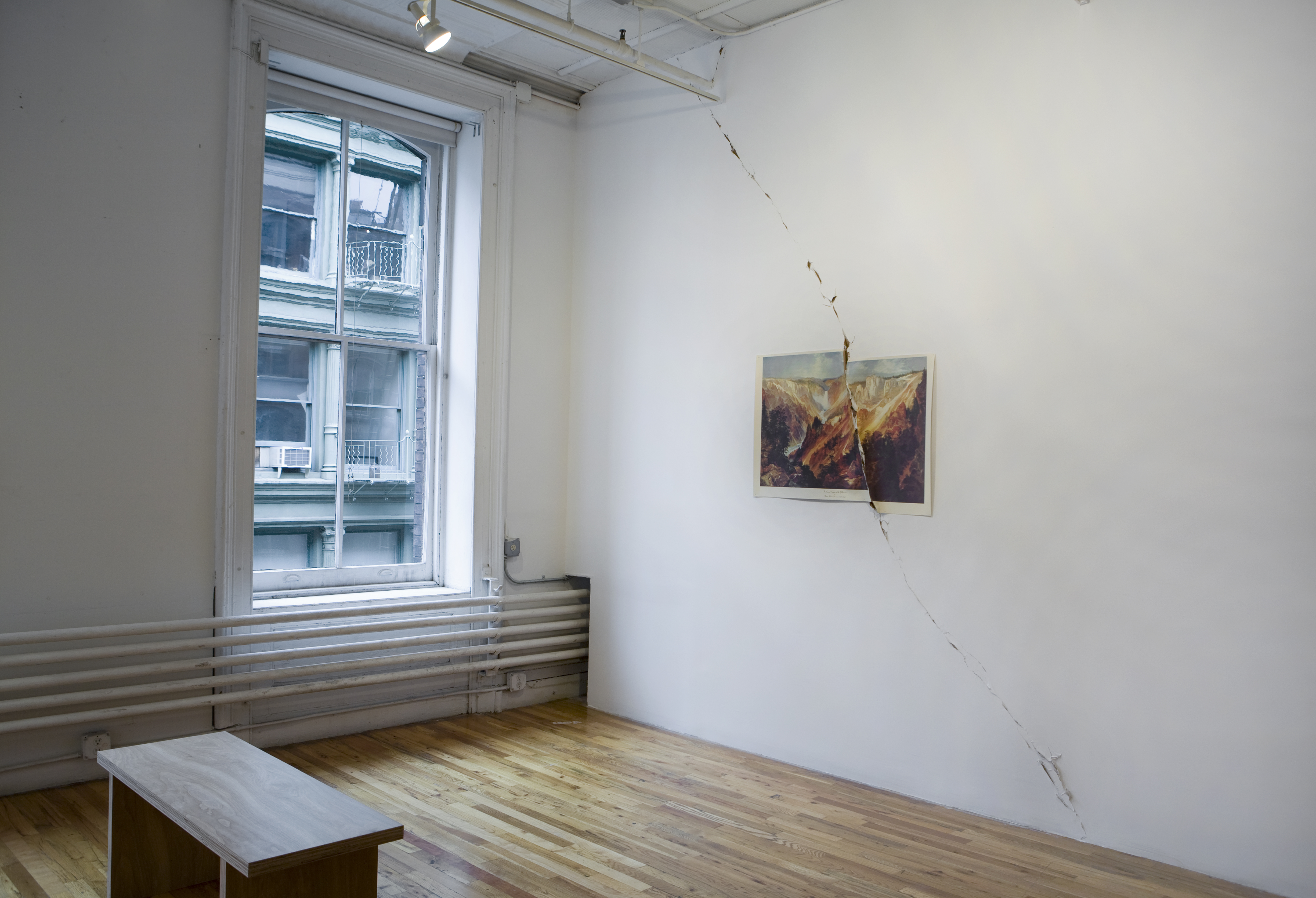 The corner of a loft space. A printed image of a canyon is affixed to the wall. The image is torn in half and the tear extends off of the paper onto the wall itself, continuing from floor to ceiling.