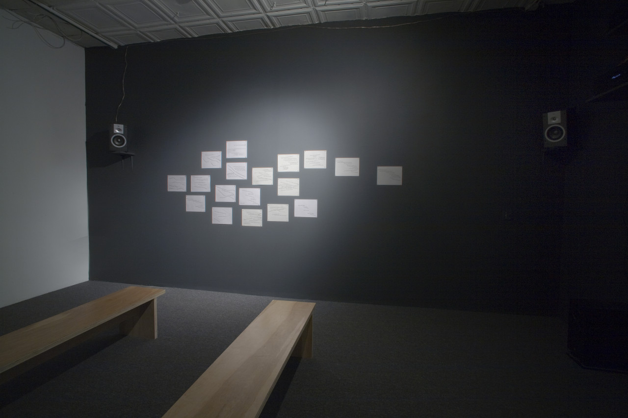 A carpeted gallery with two benches. One wall is painted dark gray and on it are two audio speakers mounted on either side. Between them are eighteen sheets of paper attached to the wall and lit with a spotlight.