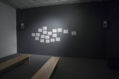A carpeted gallery with two benches. One wall is painted dark gray and on it are two audio speakers mounted on either side. Between them are eighteen sheets of paper attached to the wall and lit with a spotlight.