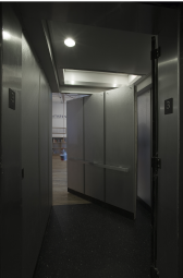 A third floor entrance off of an elevator. Beyond its black-floored vestibule is a large object standing at an angle inside the doorway. It emulates the interior panels of an elevator. A partially obscured sign saying 
