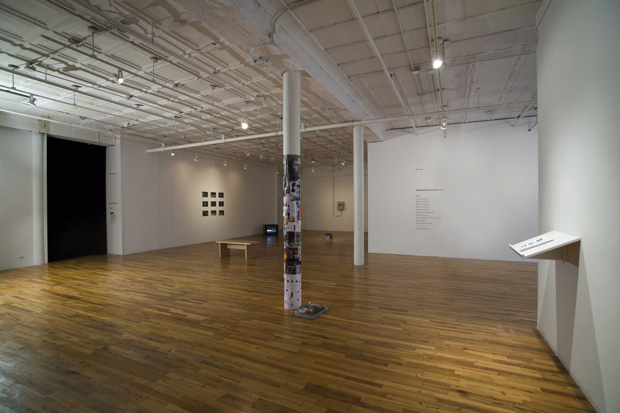 The interior of a Soho loft space. Its central column is collaged with photographs and printed paper with a small stack of materials on the floor next to it. On the left is a wide dark doorway to another room. In the distance is a bench, images on the wall in a grid, and a TV on the floor. There is wall text to the right and a wall shelf with photocopied papers on it.
