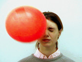 A figure with long brown hair wearing a gray sweater over a light pink and white button-down shirt clenches their eyes shut as a red ball hits their face.