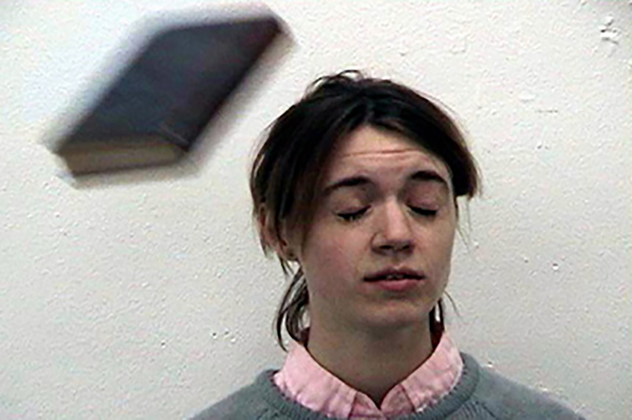 A figure with long brown hair wearing a gray sweater over a light pink and white button-down shirt closes their eyes as a thrown book flies towards their face.